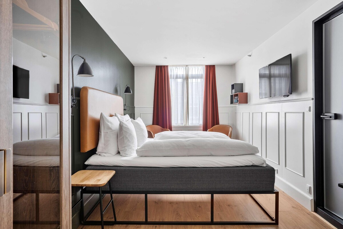 Een moderne hotelkamer met een bed, nachtkastje, lamp, televisie en een raam met gordijnen.