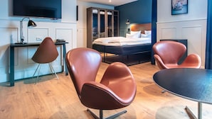 A modern hotel room with a bed, a desk with a computer, a TV, and two leather chairs.