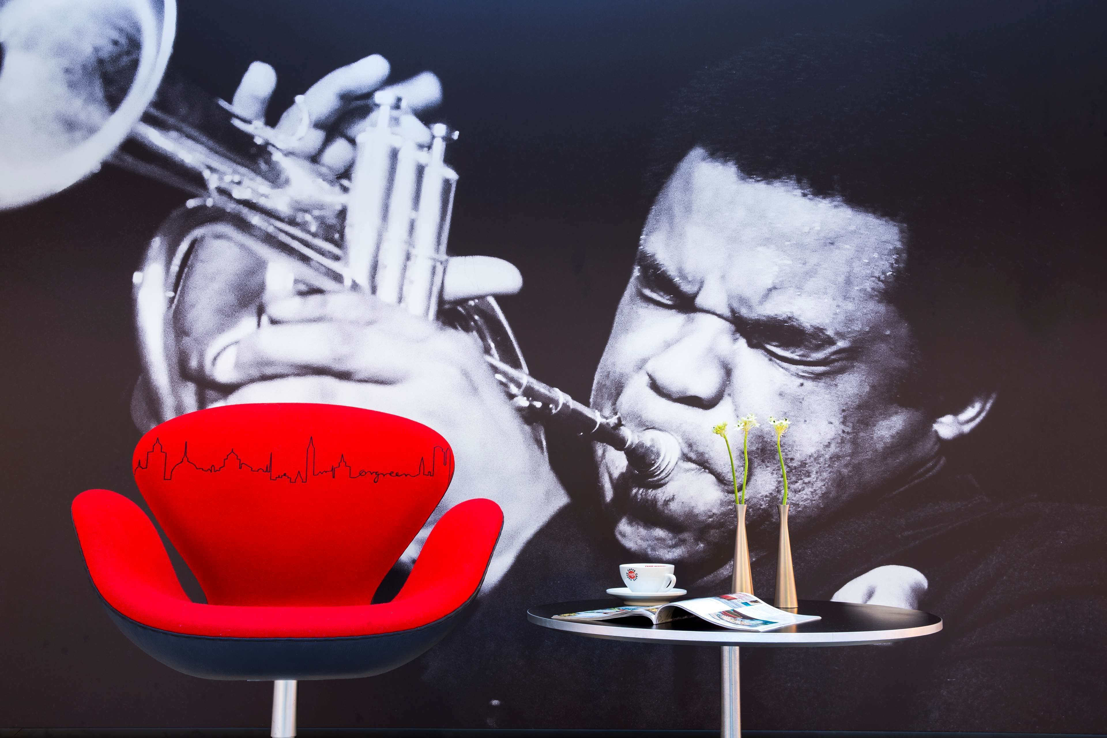 Una silla roja, una mesa con una taza y jarrones, y un cartel grande de una persona tocando una trompeta.