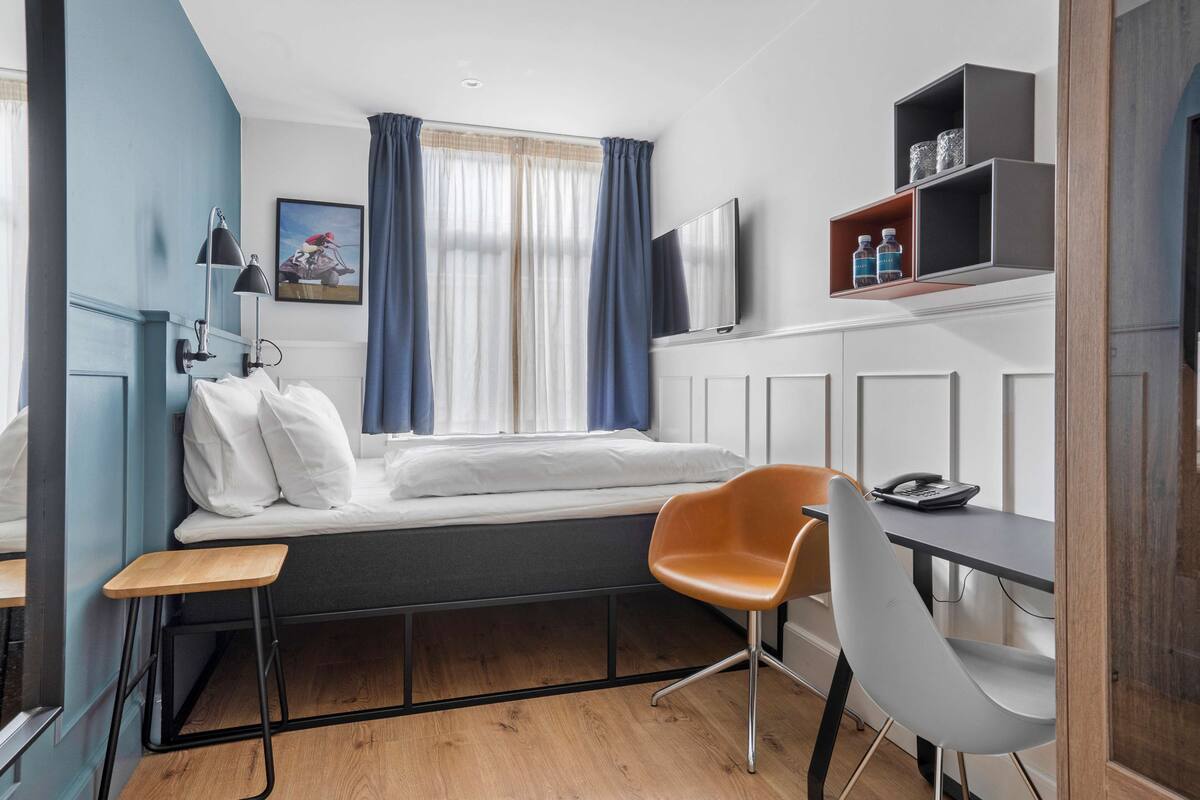 A compact room with a bed, a desk, a chair, a TV, and a shelf with bottles.