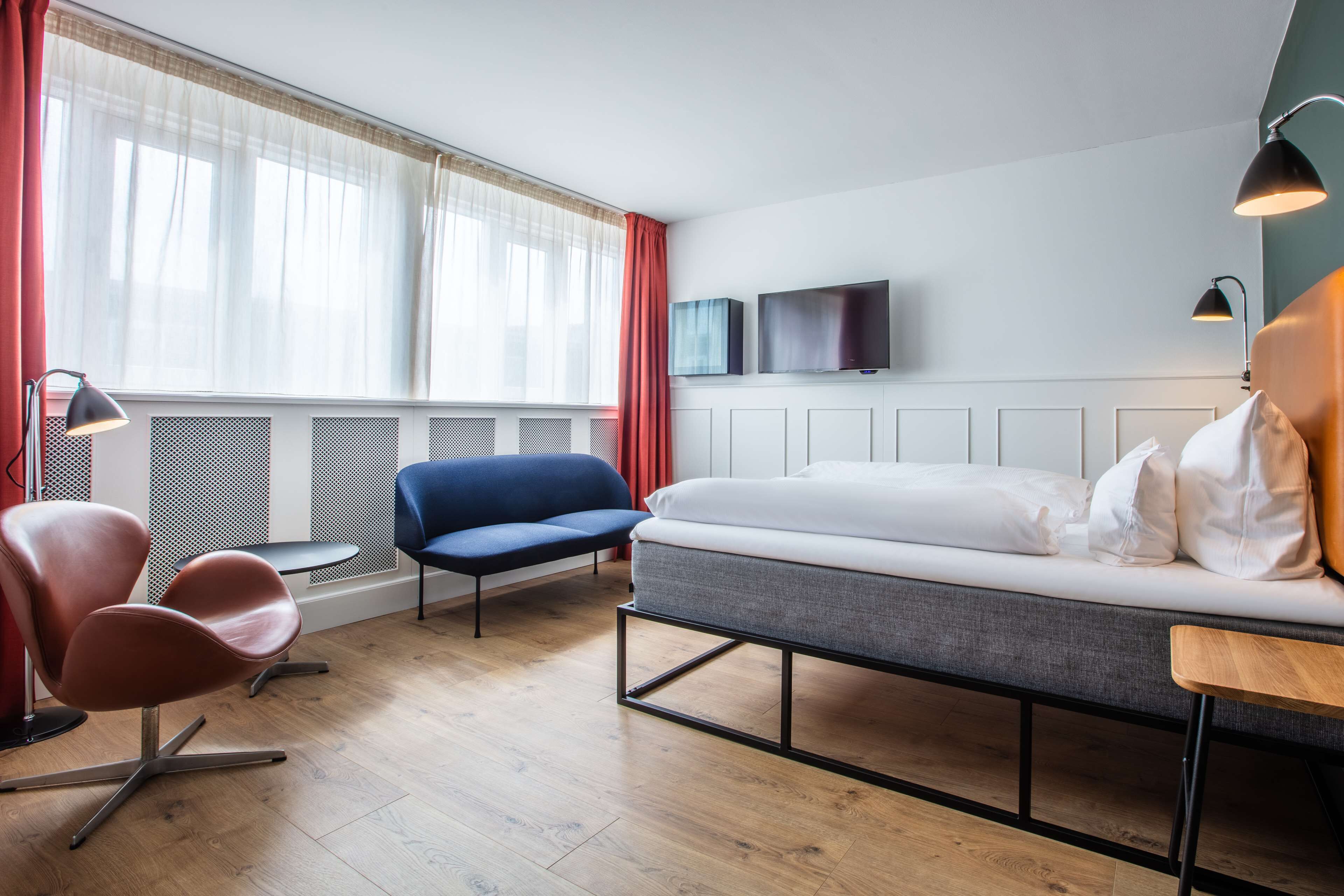 Una habitación de hotel moderna con cama, sofá, sillón, televisión y ventana con cortinas.
