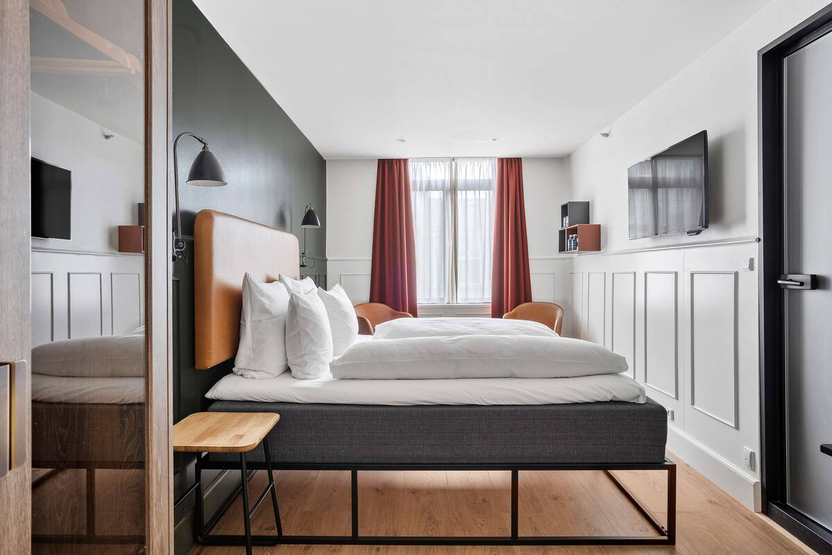 A modern hotel room with a bed, bedside table, lamp, and a television on the wall.