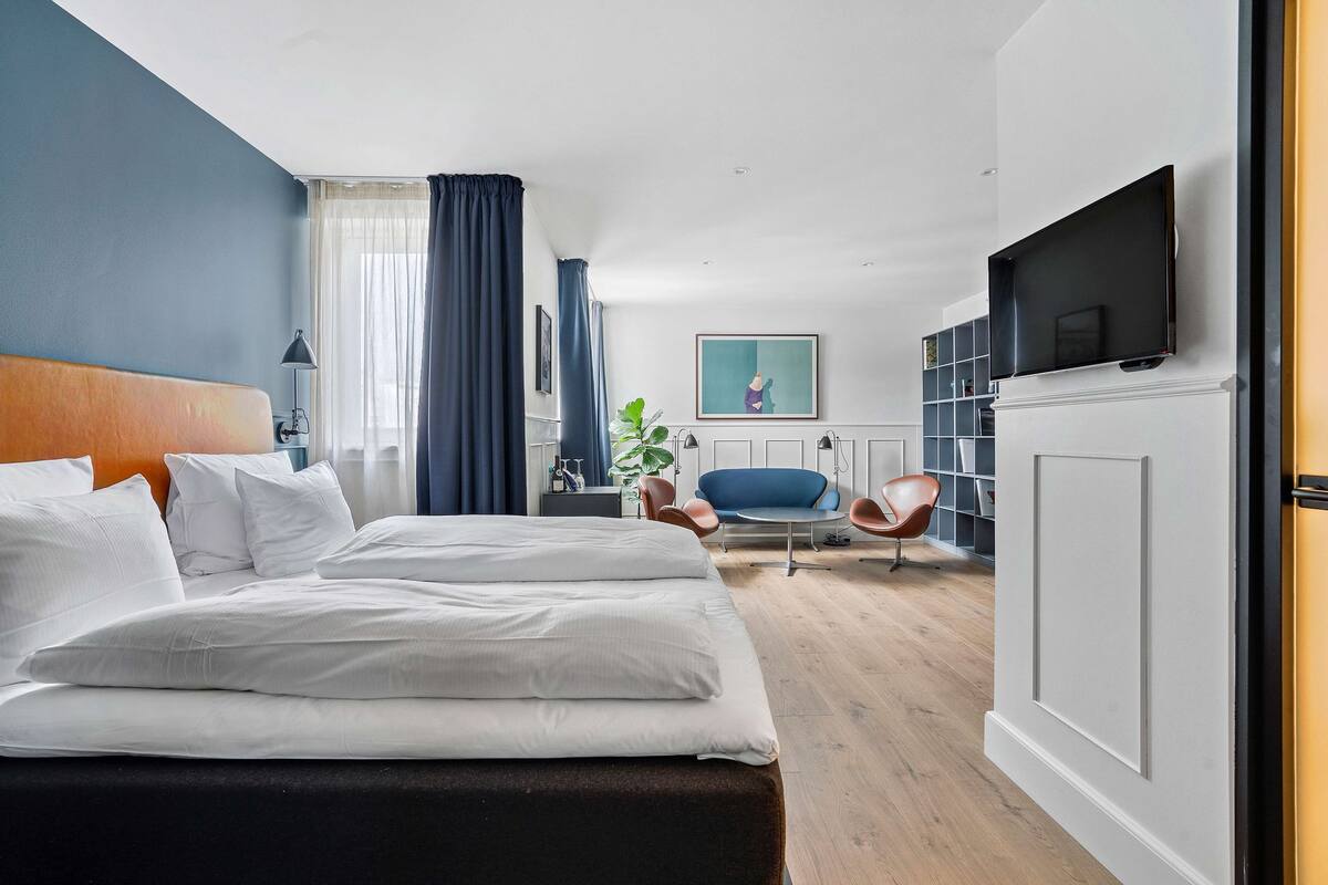 A modern hotel room with a large bed, a TV, a desk, and a seating area with chairs.