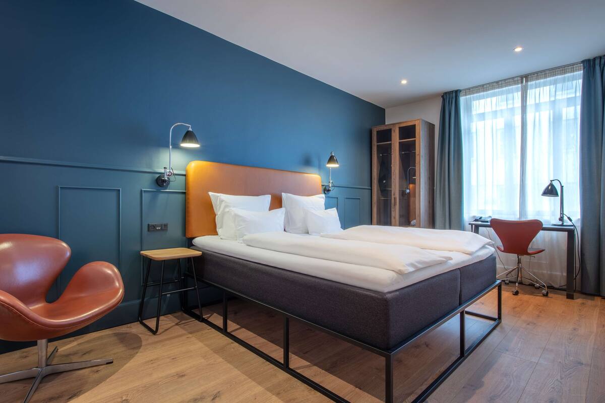 A modern hotel room with a large bed, a desk, a chair, and a wardrobe.