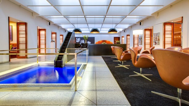 A hotel lobby with a swimming pool, seating area, reception desk, and a staircase leading to another floor.