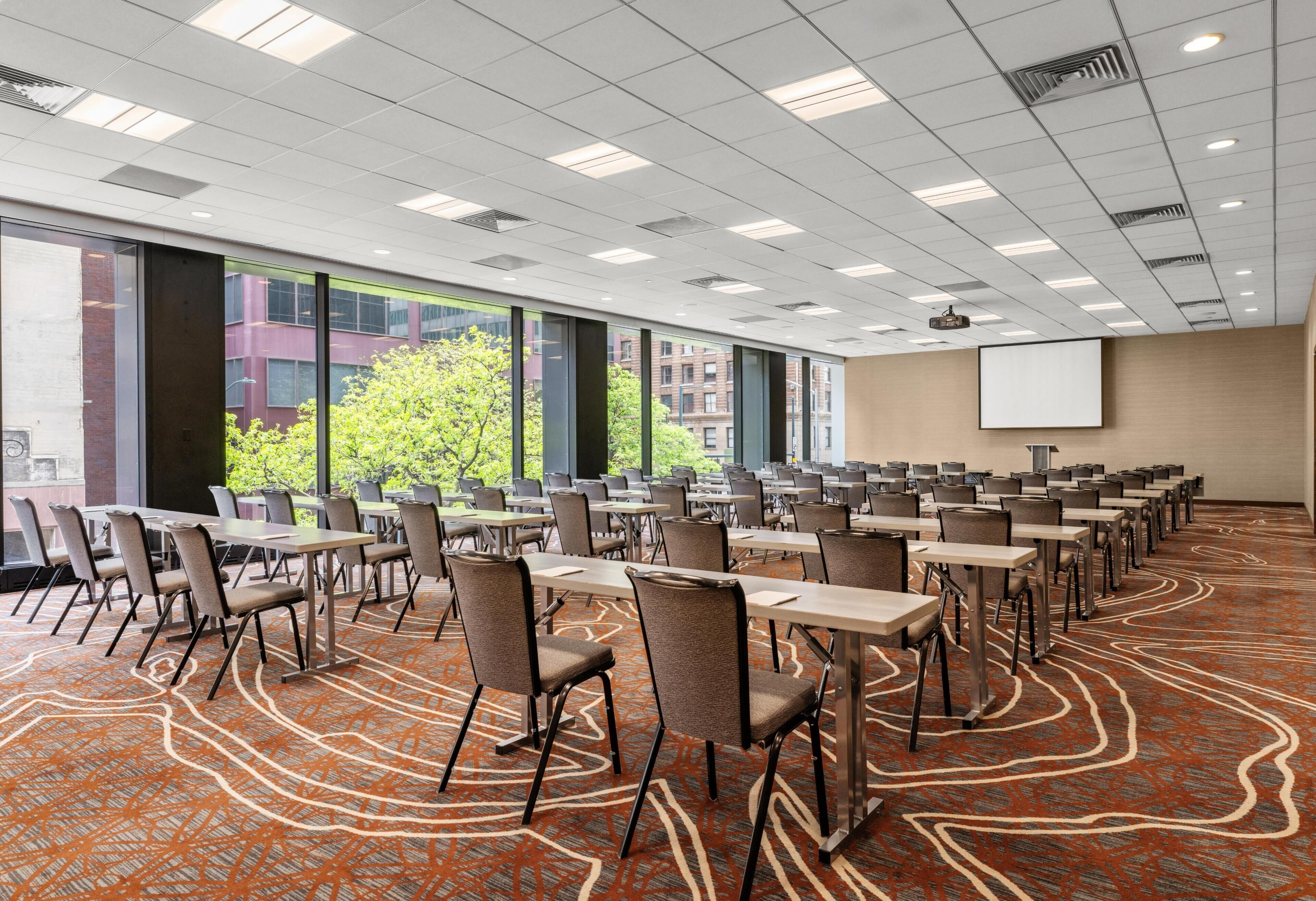 A conference room with tables and chairs, large windows, a projector screen, and a patterned carpet.