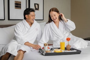 A couple in white robes on a bed with breakfast items including cereal, toast, and orange juice.