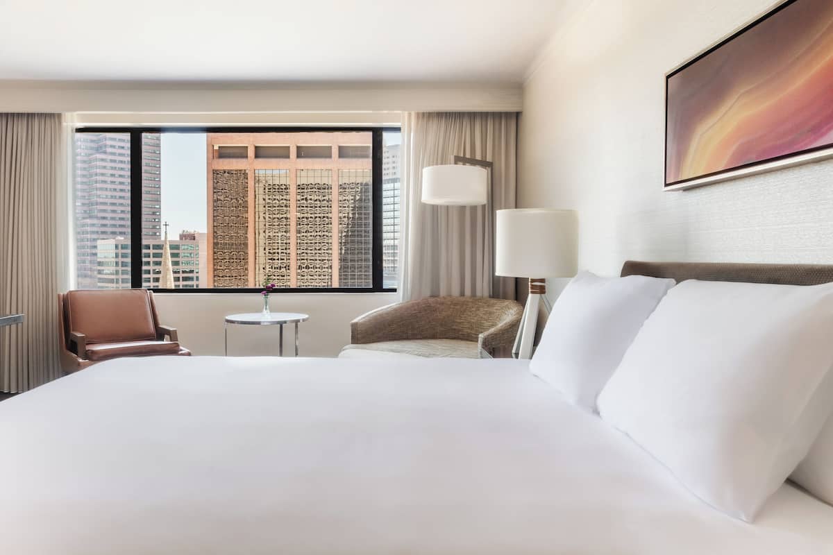 A hotel room with a large bed, two armchairs, a small table, and a large window with a city view.
