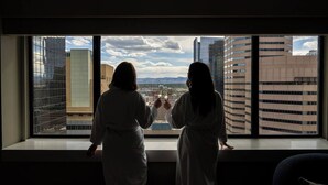 Two individuals in white robes standing by a window with a cityscape view.