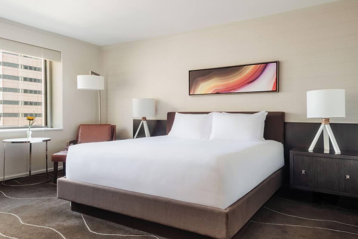 A hotel room with a large bed, two bedside tables, a chair, a desk, and a window with a view of buildings.
