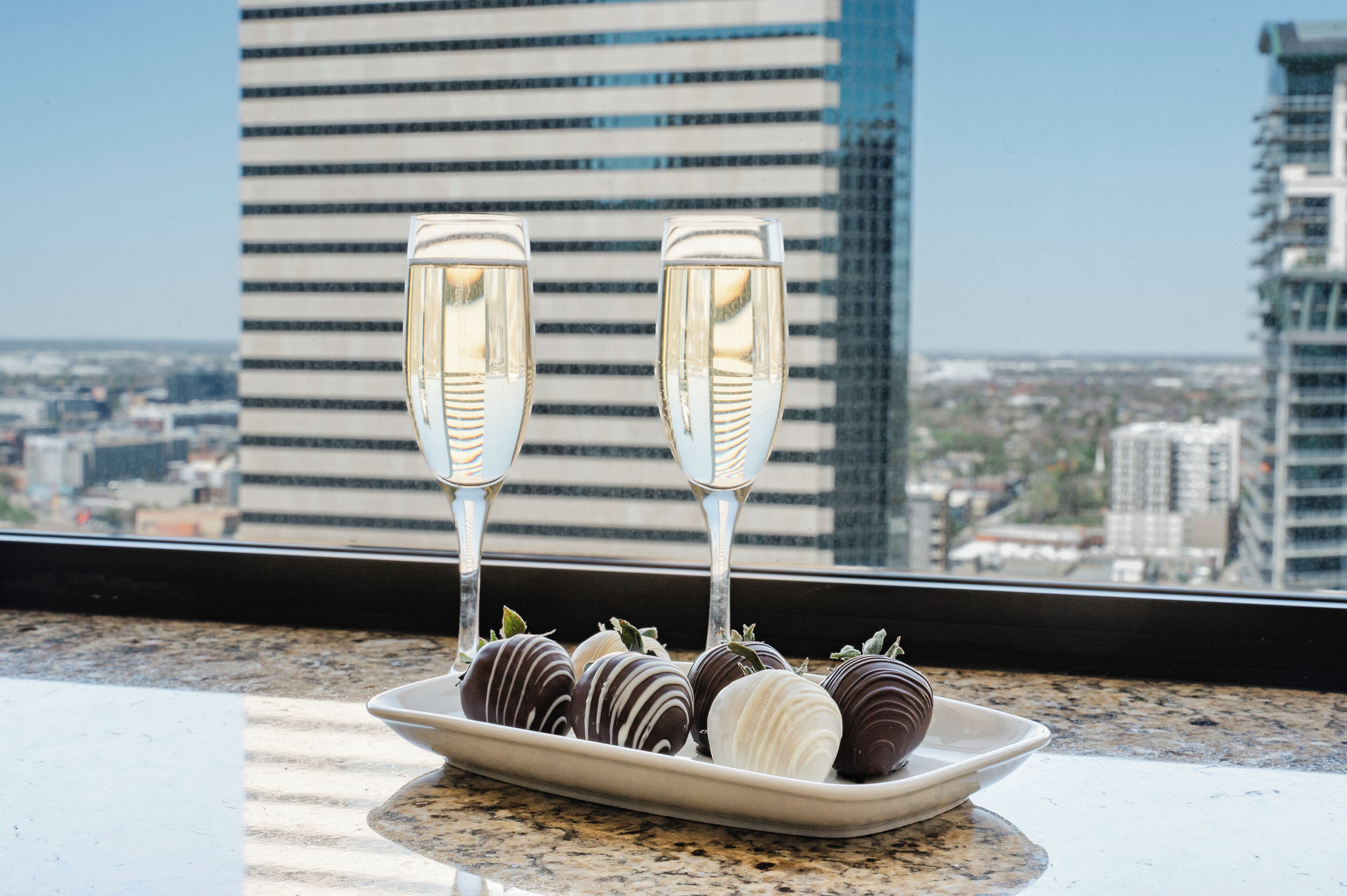 En tallerken med chokoladeovertrukne jordbær og to champagneglas på et vindueskar med udsigt over bybilledet.