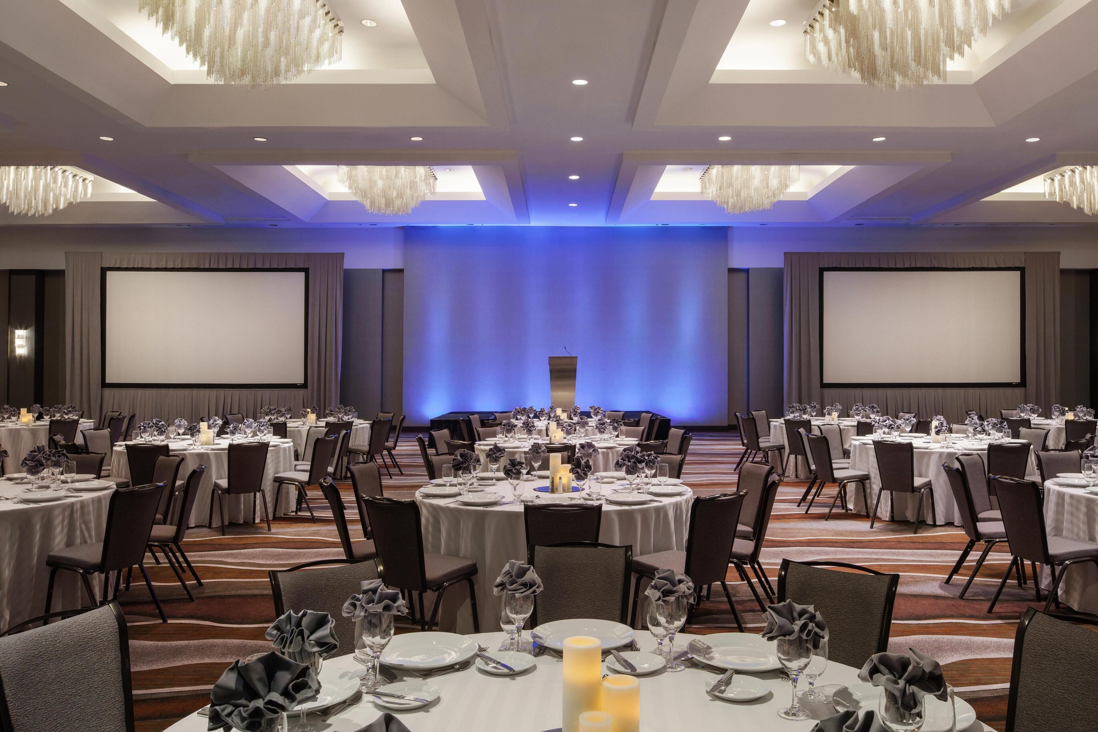 Un salón de conferencias con varias mesas dispuestas para comidas, equipado con sillas y mesas, y una pantalla grande al frente.