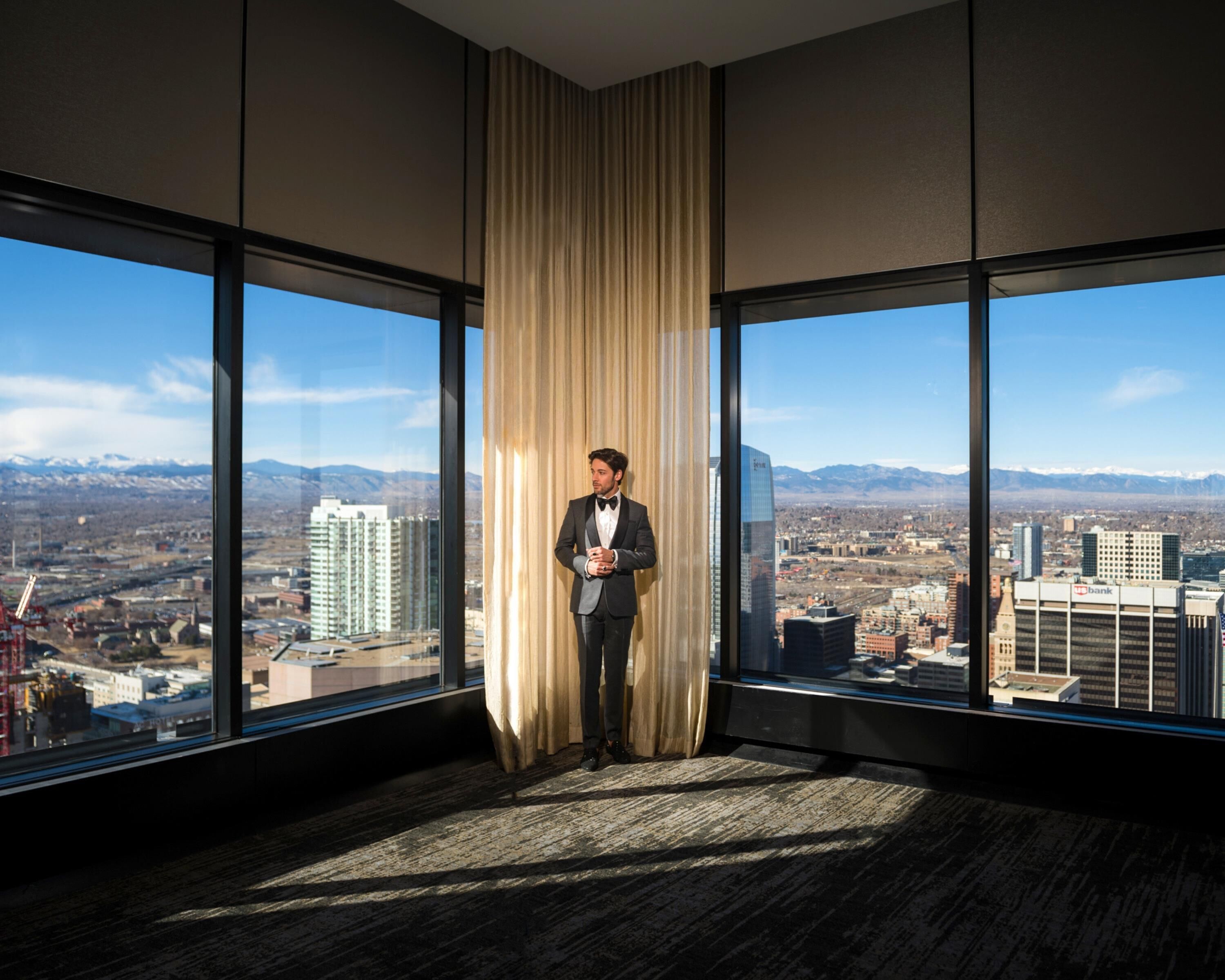 Un hombre está de pie en una habitación con ventanas amplias que dan a una vista panorámica de la ciudad y montañas.