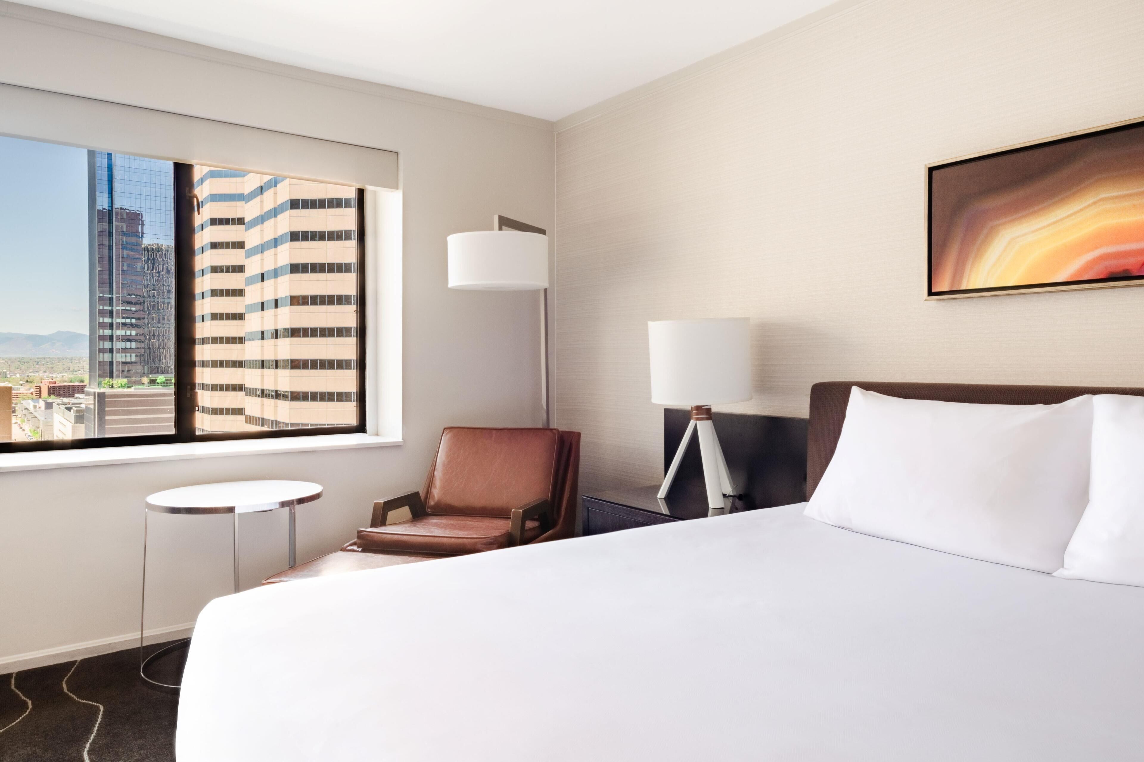 A hotel room with a large bed, a chair, a desk, a lamp, and a painting on the wall.