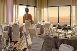 A wedding reception room with tables set for guests, large windows with a view, and a bride in the background.