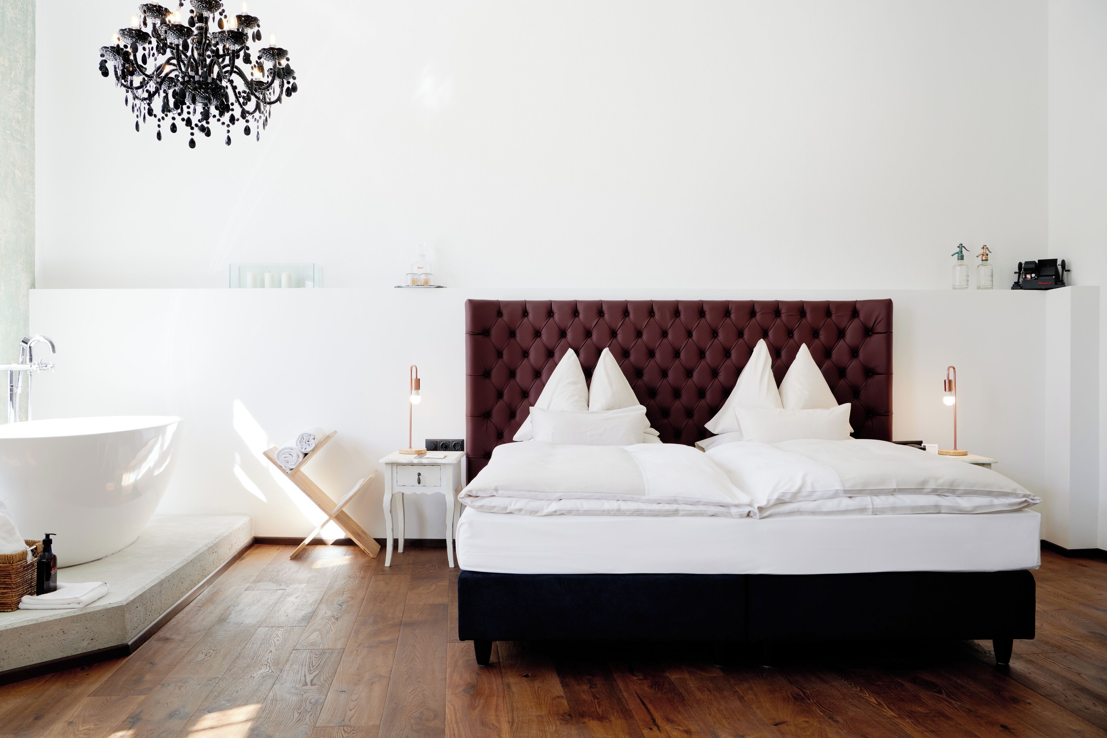 Ein modernes Schlafzimmer mit einer gepolsterten Bettseite, einem großen Bett mit weißer Bettwäsche, Holzboden, einer weißen Badewanne und einem Kronleuchter.