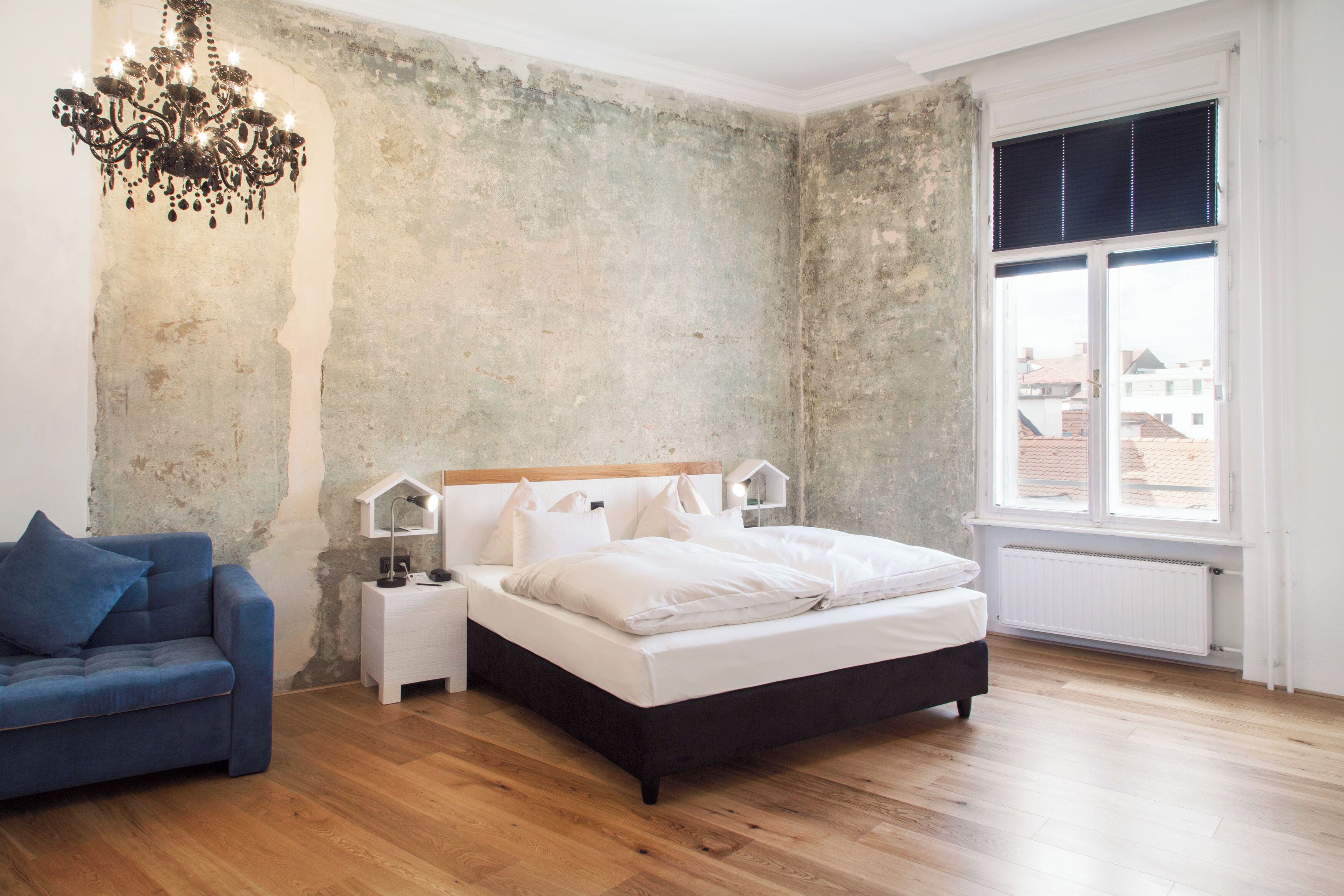 Un dormitorio con cama, mesita de noche, sofá azul, candelabro y un amplio ventanal con persianas.