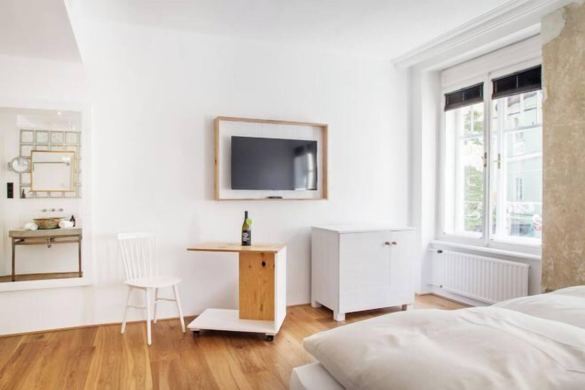 A bedroom with a bed, a desk, a chair, a TV, and a window.