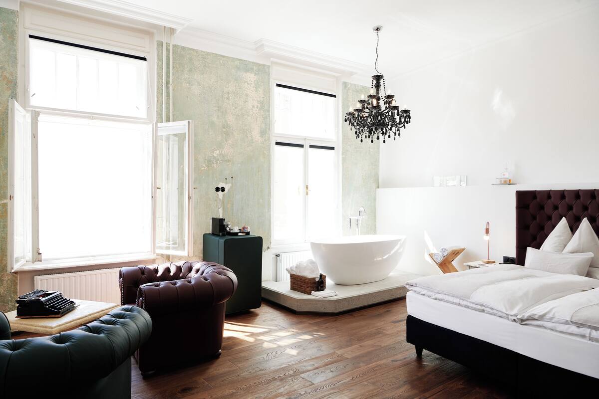 A modern bedroom with a large bed, a sitting area with a leather sofa, a bathtub, and a chandelier.