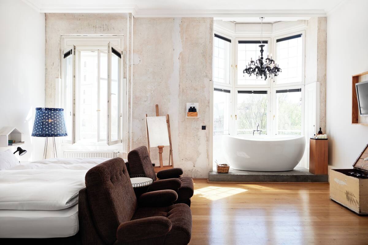 A bedroom with a large bed, a sitting area with brown sofas, a bathtub, and a chandelier.