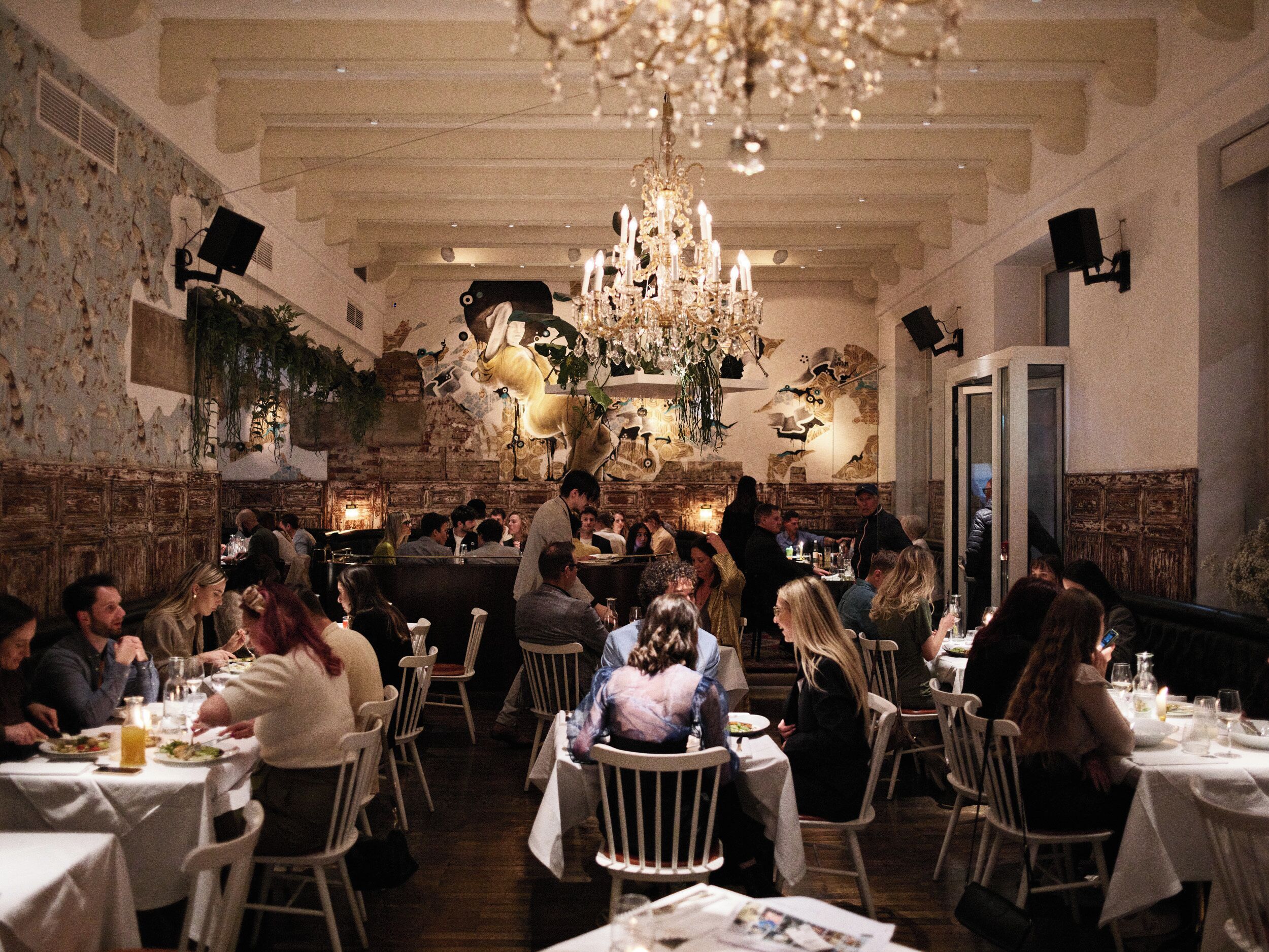 Een restaurant met witte tafels en stoelen, een kroonluchter en een bakstenen muur.