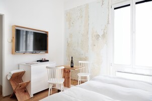 Habitación minimalista con una cama, un escritorio, dos sillas, un televisor y una ventana.