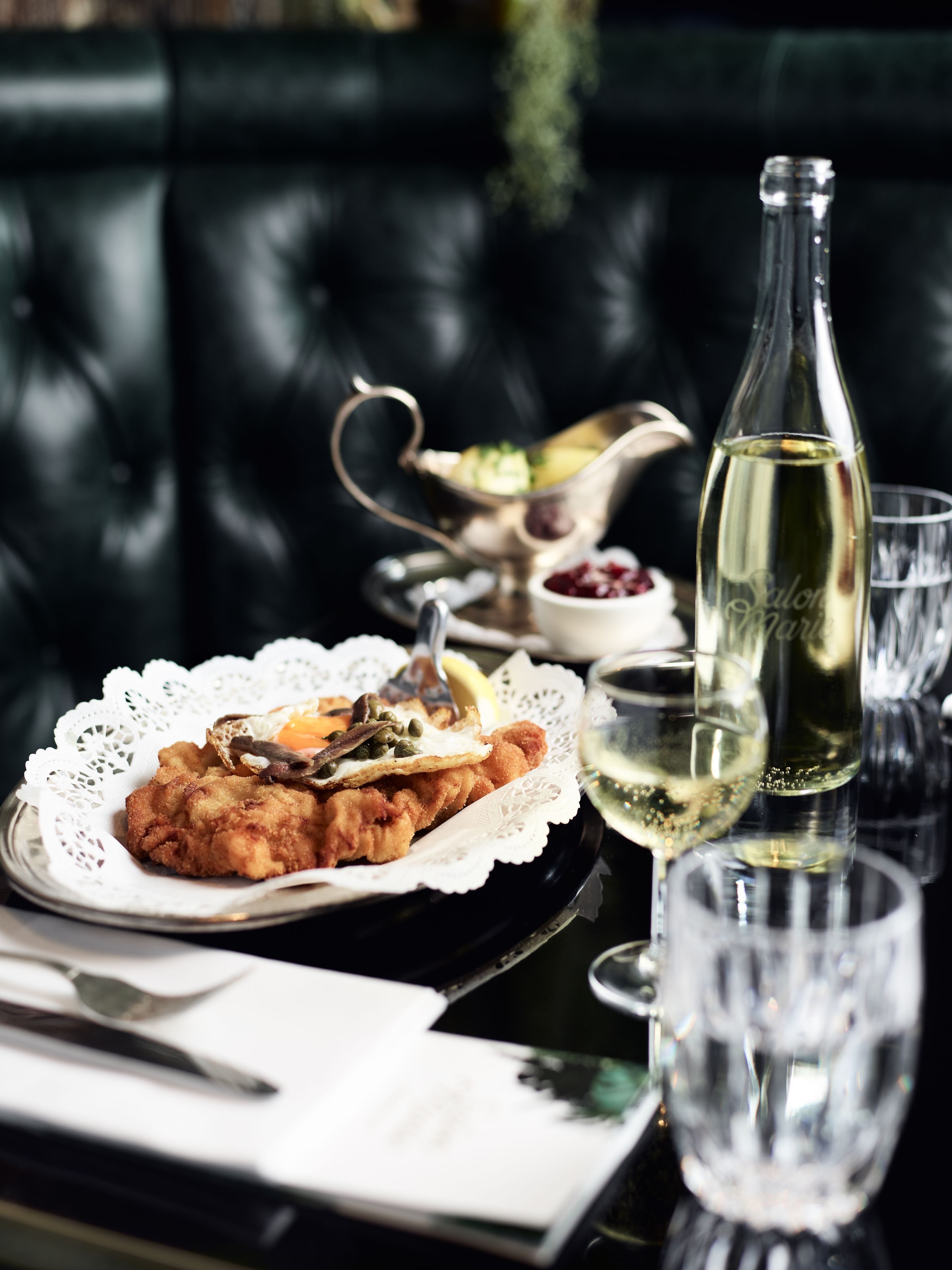 A dining table with a plate of fried food, a bottle of white wine, and a silver gravy boat.
