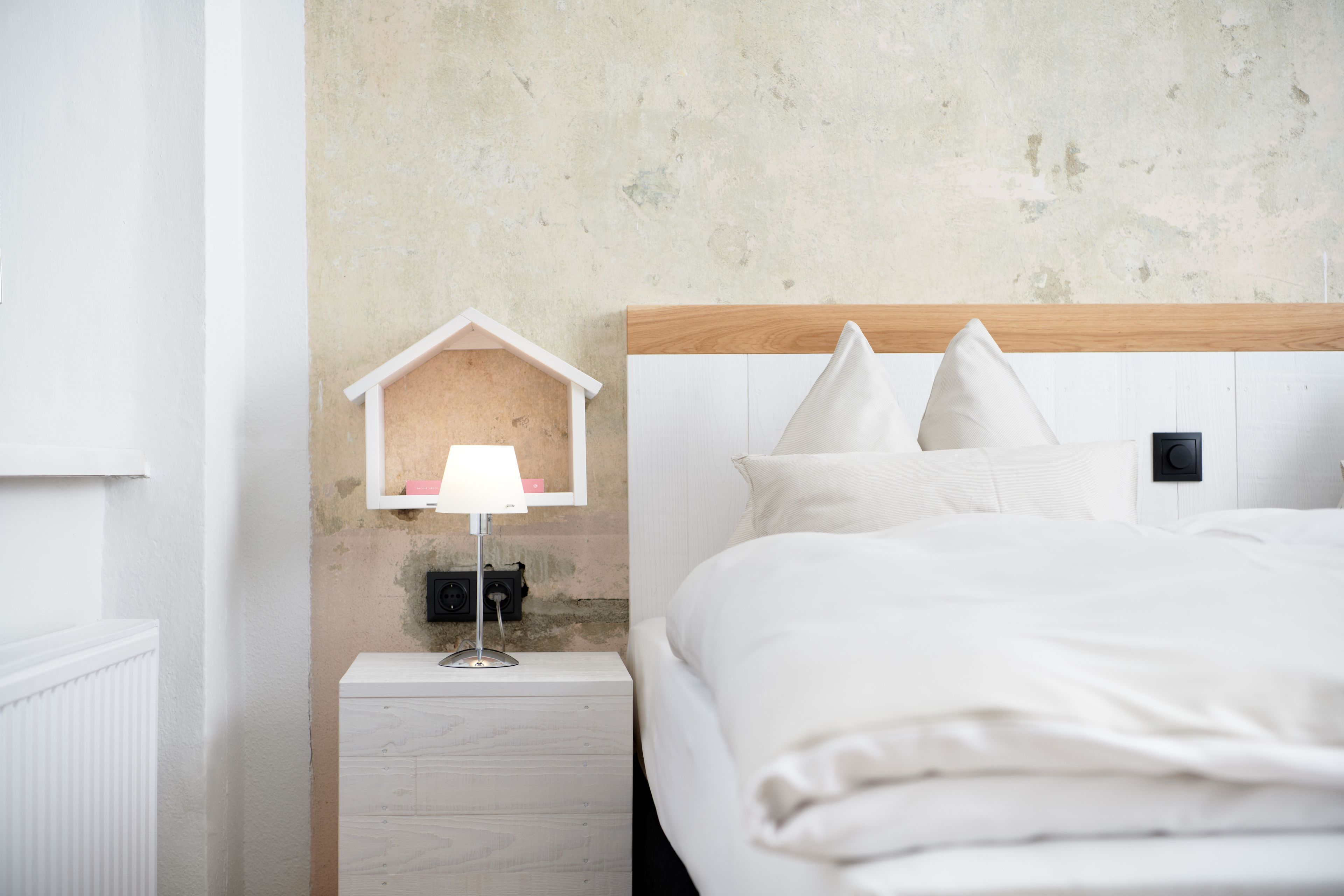 A neatly made bed with white bedding, a bedside lamp, and a small wooden house-shaped shelf with a lamp on it.