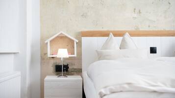 A neatly made bed with white bedding, a bedside lamp, and a small wooden house-shaped shelf with a lamp on it.