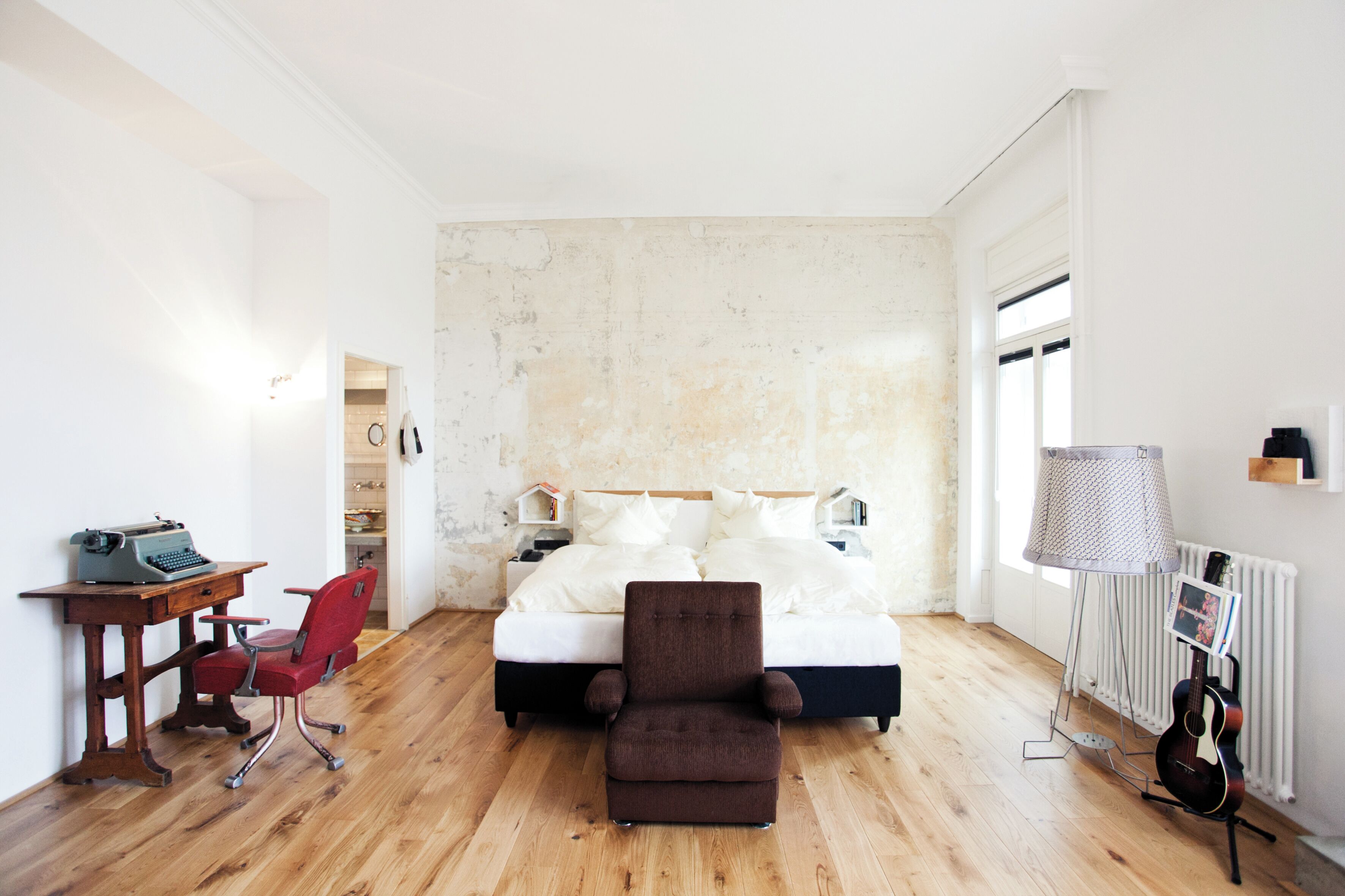 A bedroom with a bed, a desk with a typewriter, a red chair, a brown armchair, a guitar, and a radiator.