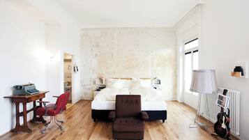 A bedroom with a bed, a desk with a typewriter, a red chair, a brown armchair, a guitar, and a radiator.