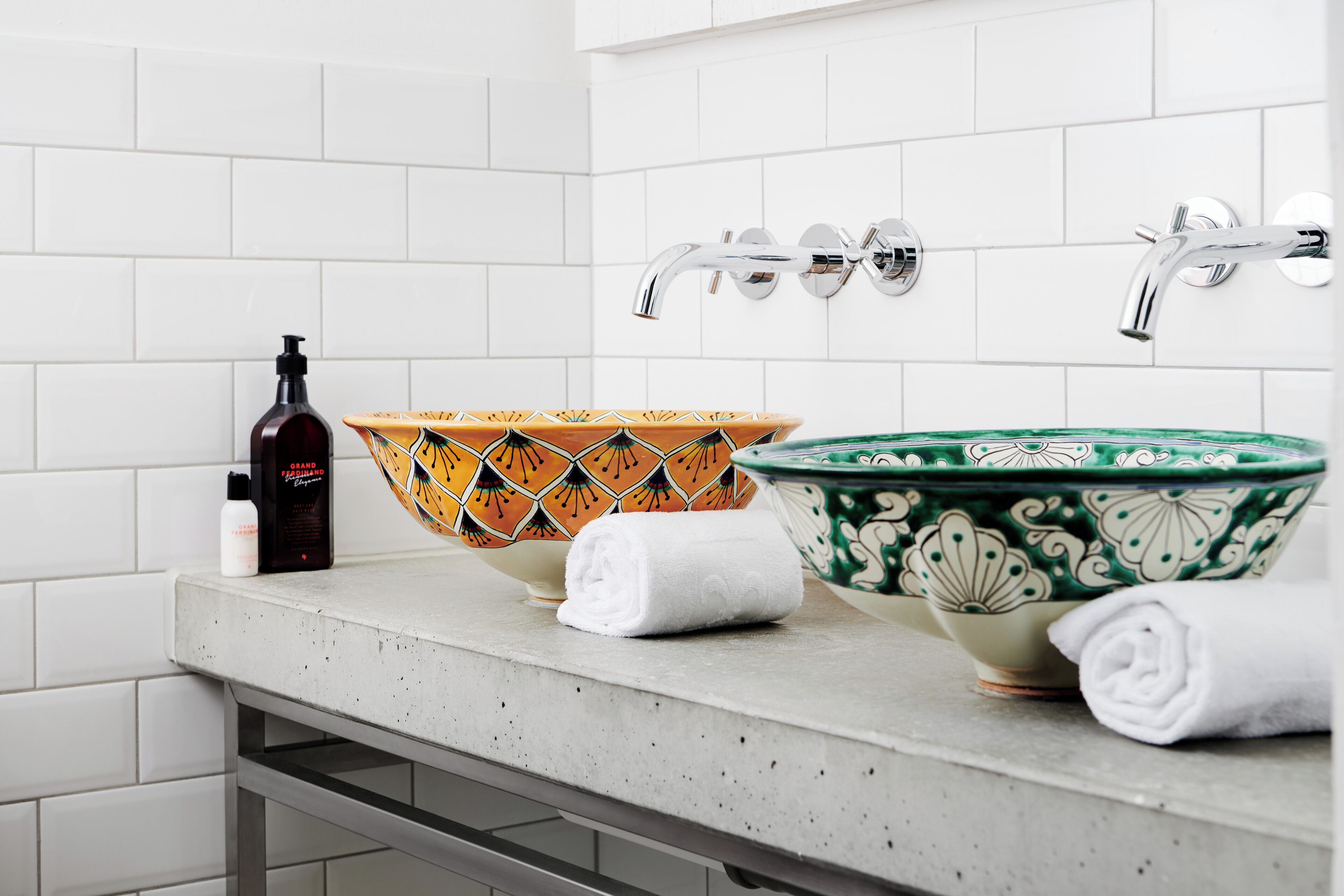A bathroom with white tiled walls, a countertop with two bowls, a bottle, and two towels.