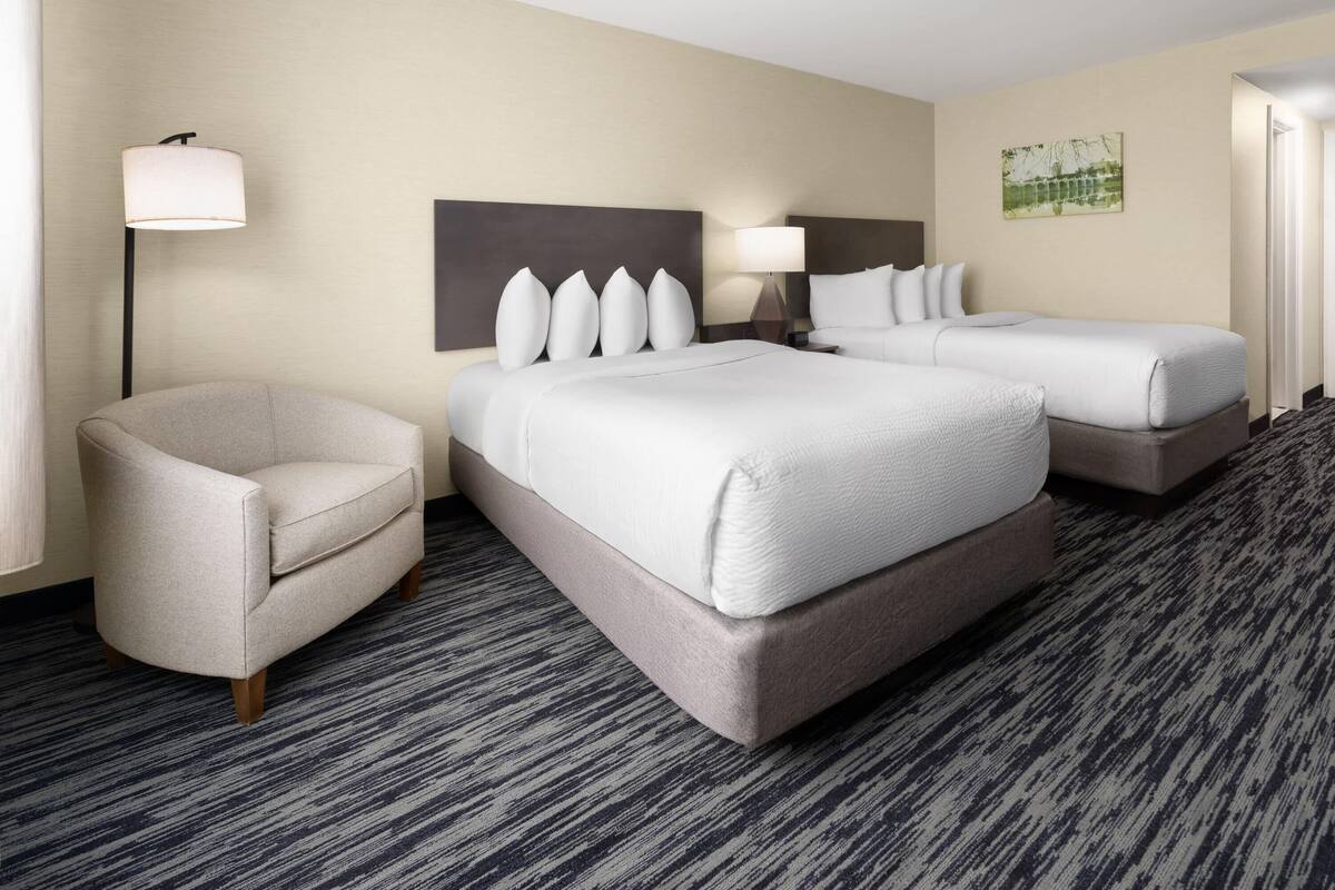 A hotel room with two beds, a chair, and a lamp.