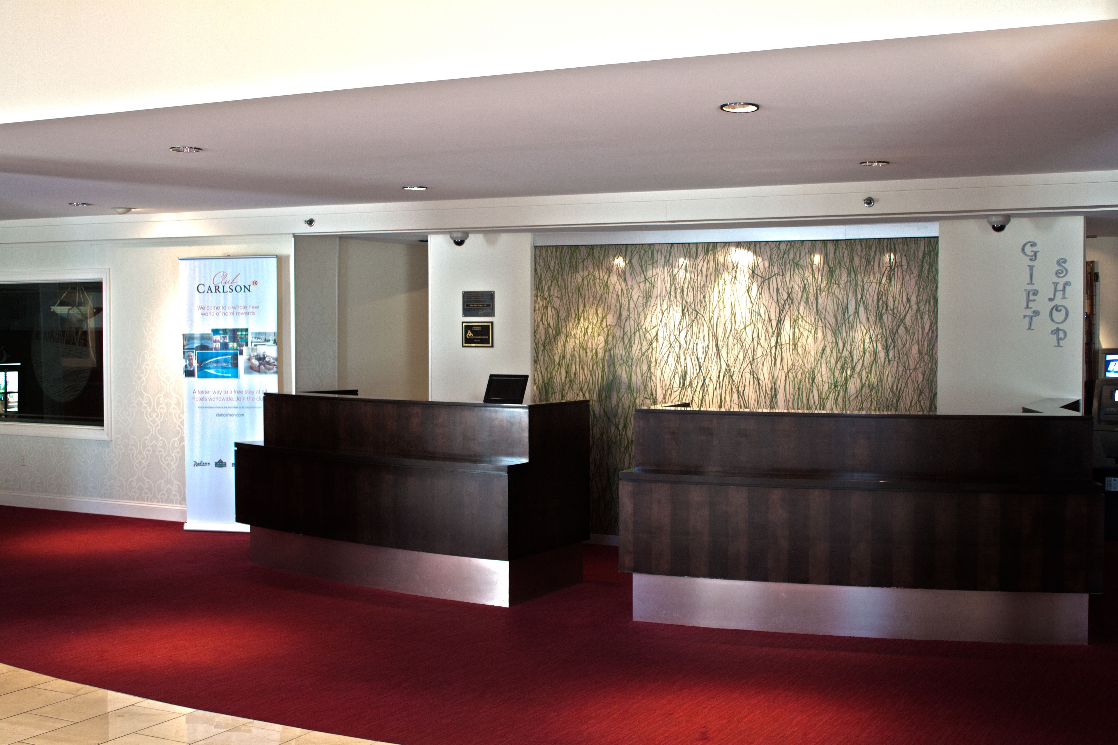 Un hall d’hôtel comprenant un comptoir d’accueil avec deux postes de réception, un tapis rouge, un mur agrémenté d’un jardin vertical et un panneau indiquant l’emplacement de la boutique de souvenirs.