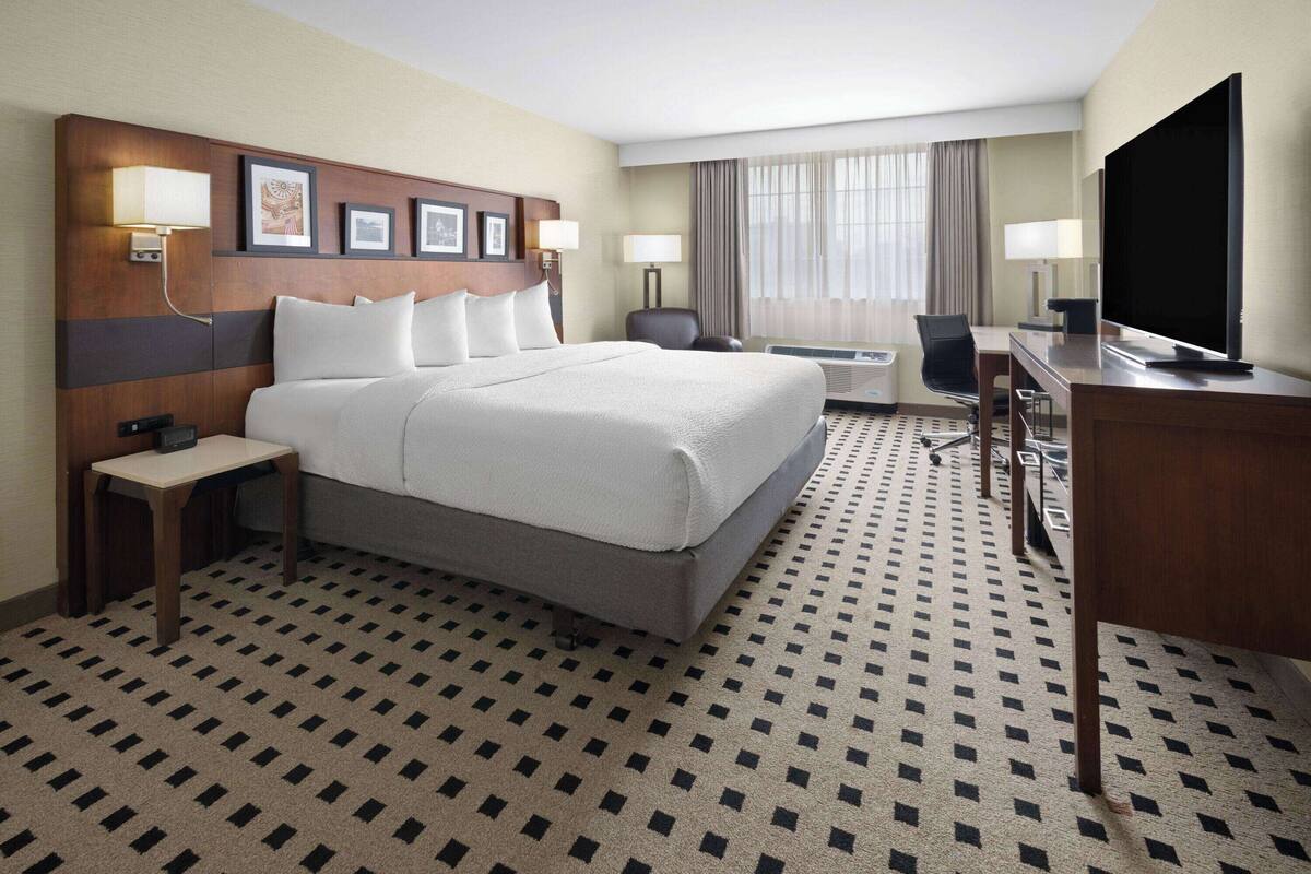 A hotel room with a large bed, a desk with a flat-screen TV, a chair, and a desk lamp.