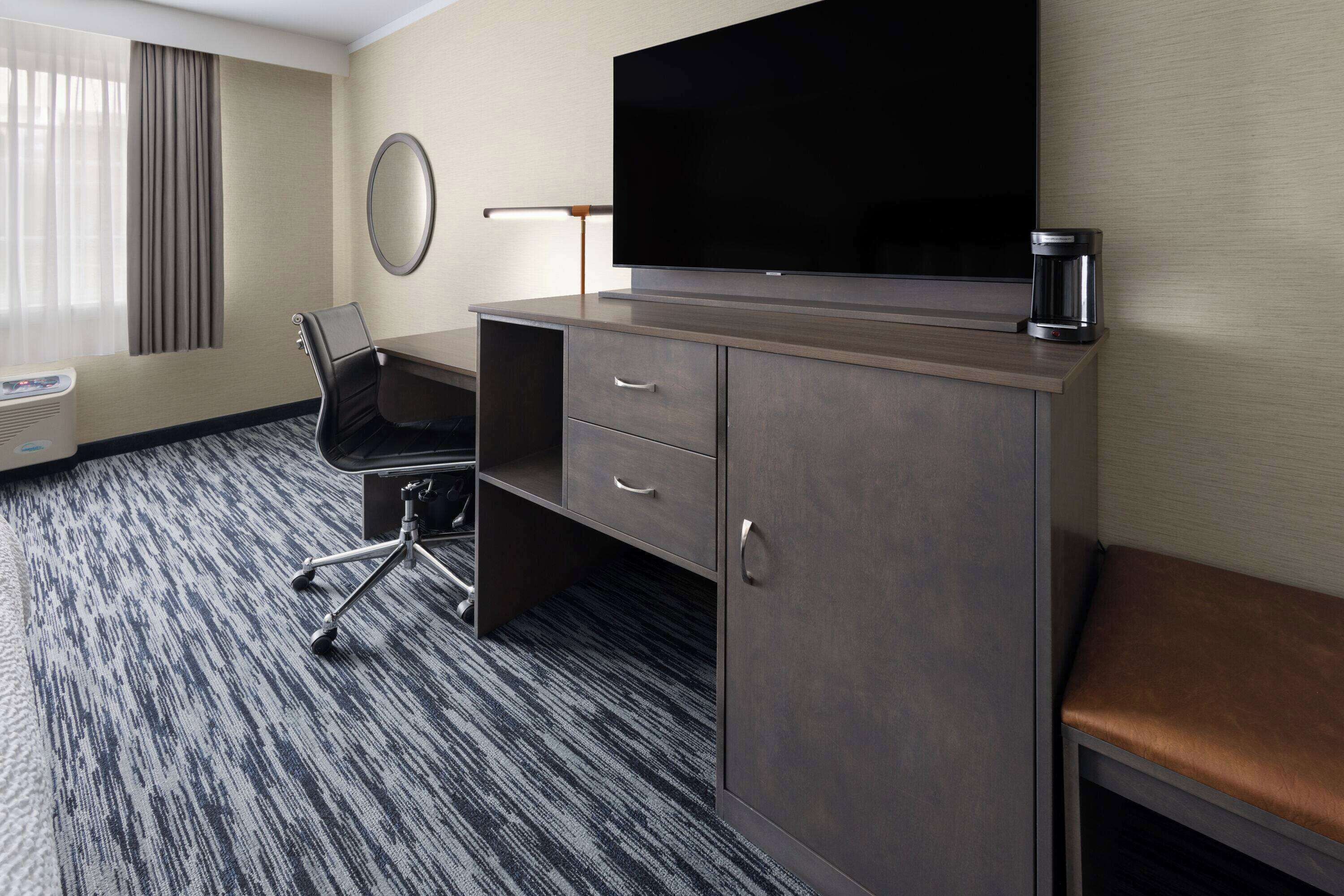 A hotel room with a large flat-screen TV, a desk with drawers, a chair, a coffee maker, and a bench.