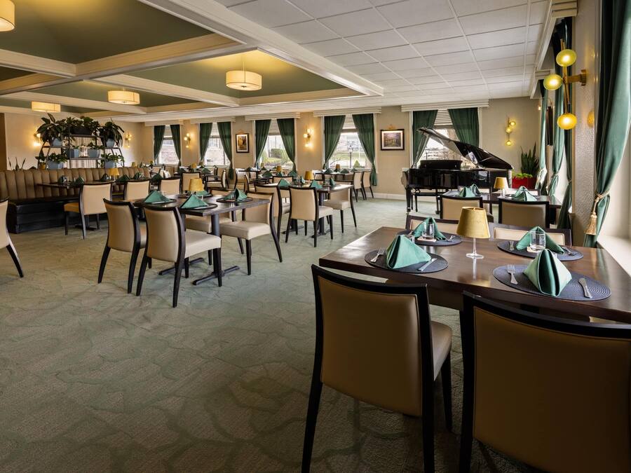A spacious dining area with multiple tables and chairs, a piano, and a bar area with plants and framed pictures.