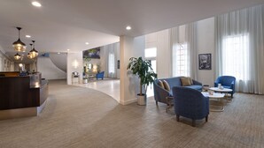Eine großzügige Hotelhalle mit einer Rezeption, einer Sitzecke mit Sofas und Sesseln, einer großen Topfpflanze und einer modernen Treppe.