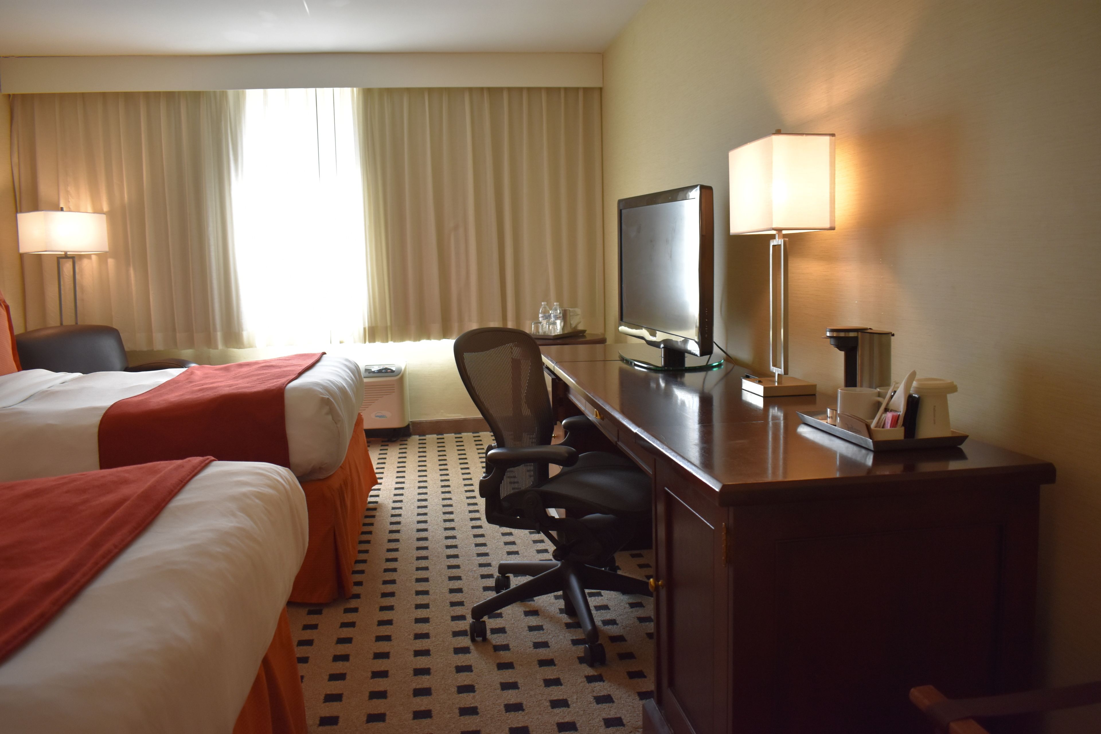 Habitación de hotel con dos camas, un escritorio con computadora, una silla, una lámpara y un ventanal con cortinas.