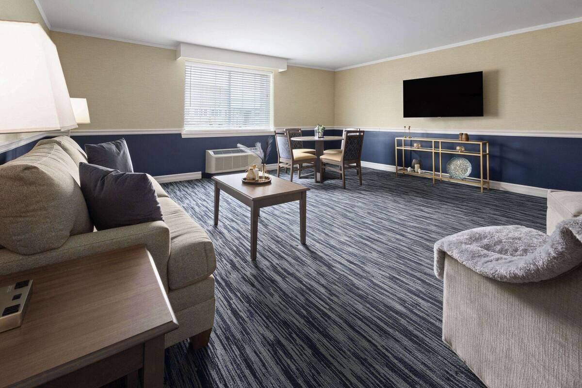 A hotel room with a sofa, a table, a TV mounted on the wall, and a window with blinds.