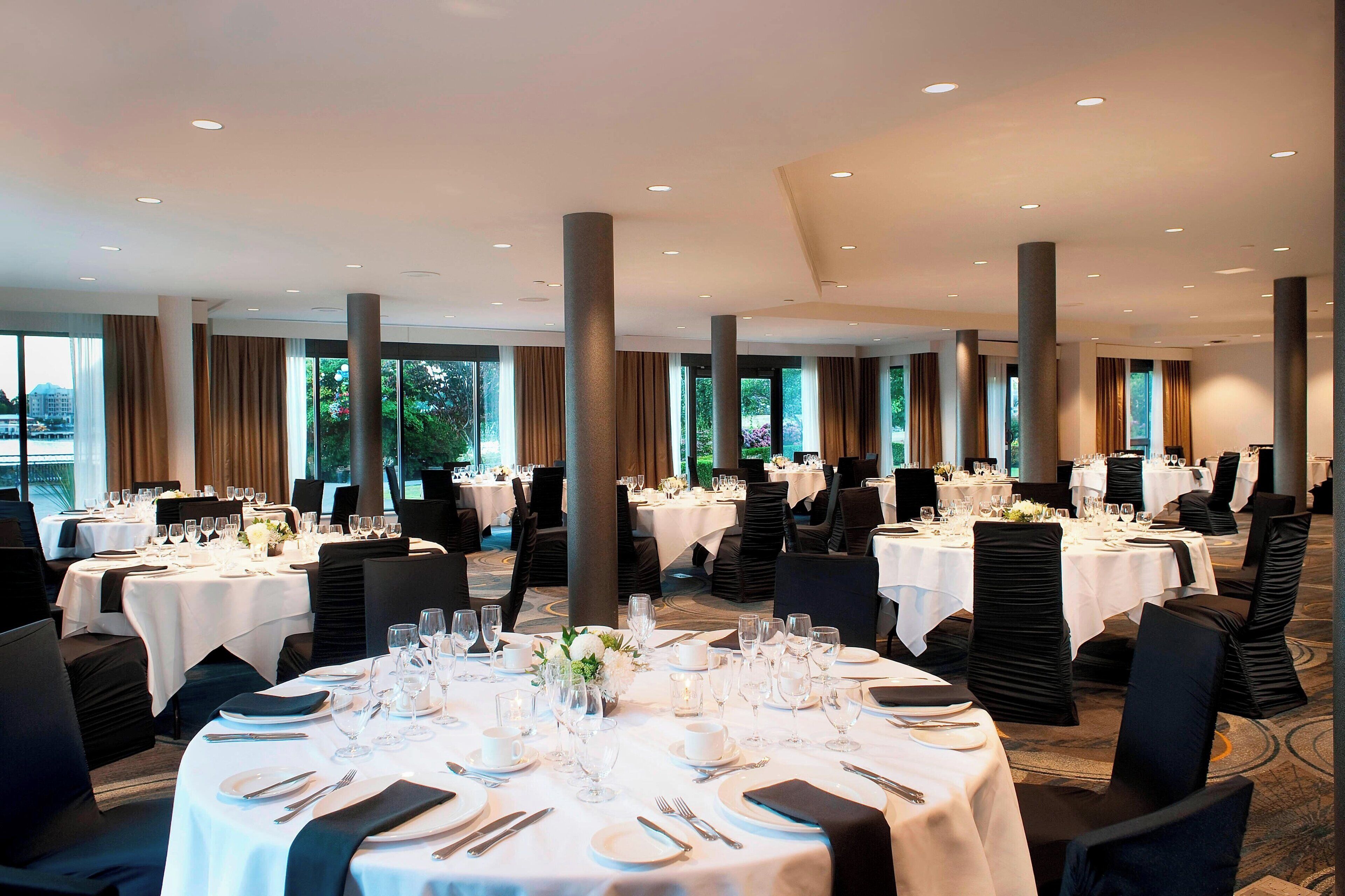 A banquet hall with round tables set for dining, large windows, and a high ceiling with recessed lighting.