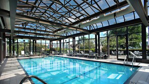 An indoor swimming pool with clear glass walls, sunlit by natural light, and surrounded by lounge chairs and tables. - Delta Hotels by Marriott Victoria Ocean Pointe Resort (Victoria)