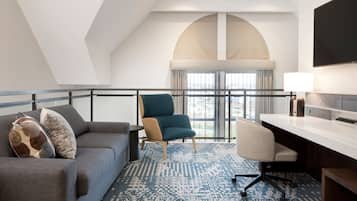 A modern hotel room with a grey sofa, a teal armchair, a desk, and a large window with a view.