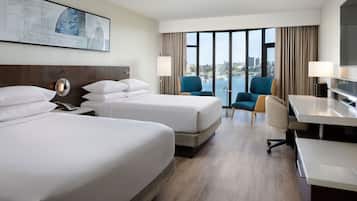 A hotel room with two beds, a desk, a chair, and a large window offering a view of the city.