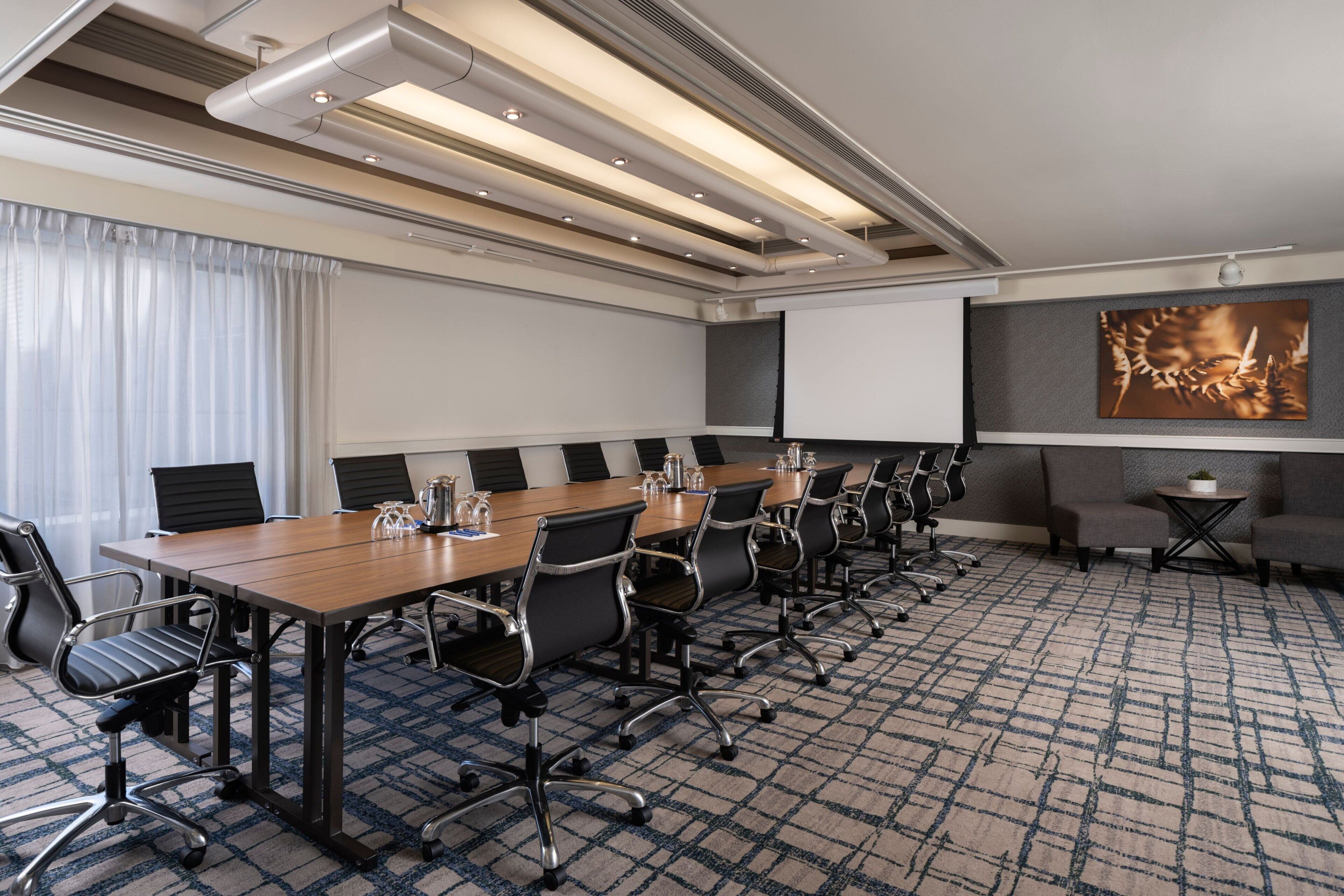 A conference room with a long table, chairs, a projector screen, and a large painting on the wall.