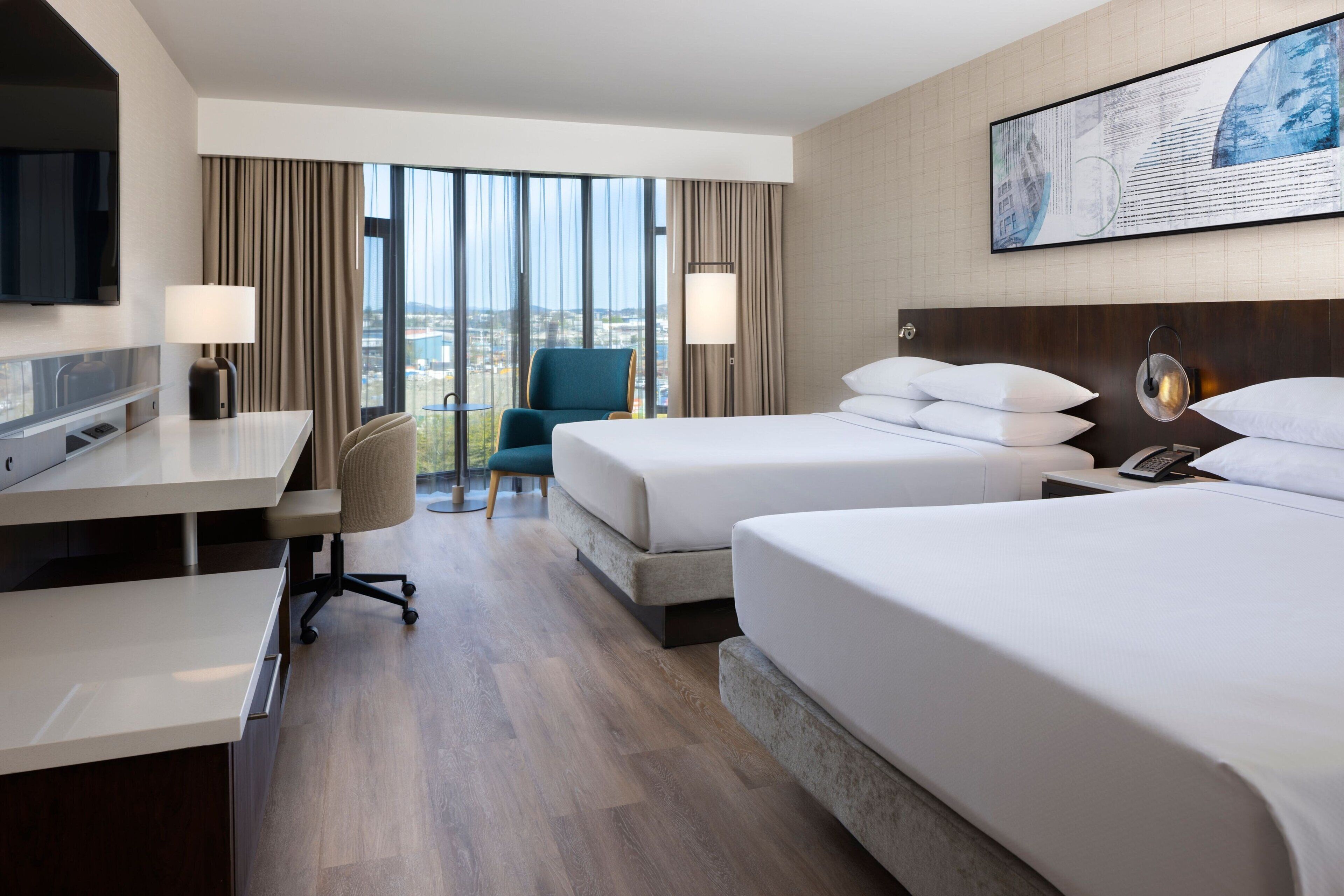 A hotel room with two beds, a desk, a chair, and a large window with a view.