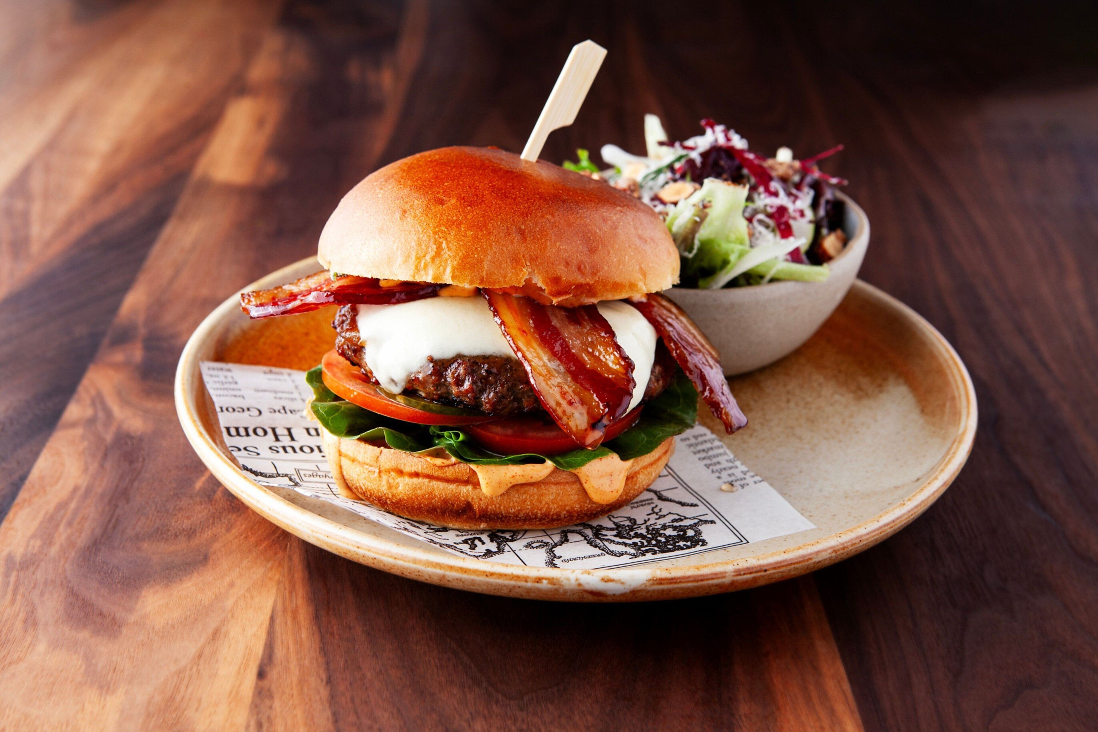 A burger with bacon, lettuce, tomato, and a bun on a plate, accompanied by a side salad.