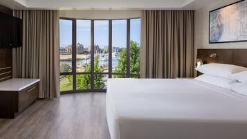 A hotel room with a large bed, a TV, a desk, and a view of a cityscape through large windows.