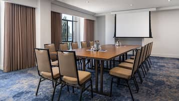 A conference room with a long table, multiple chairs, a projector screen, and large windows with curtains.