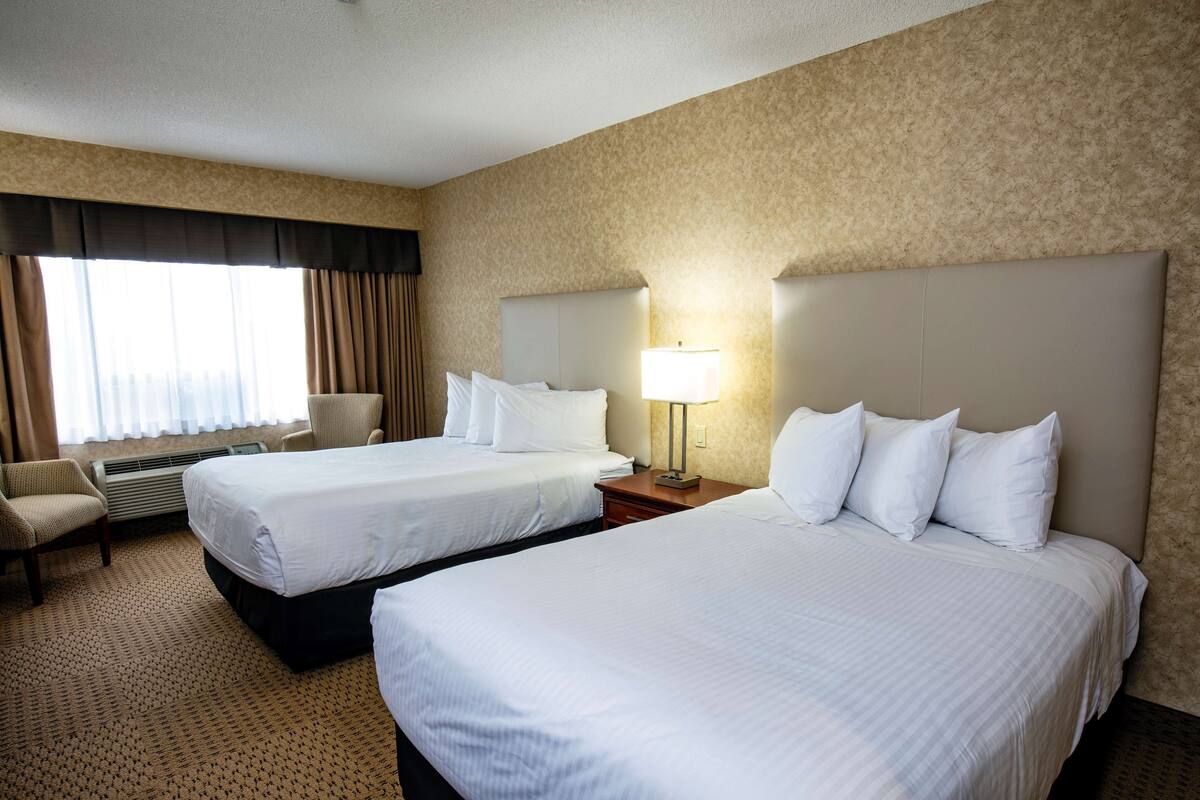 Habitación de hotel con dos camas, una silla, una lámpara y un amplio ventanal con cortinas.
